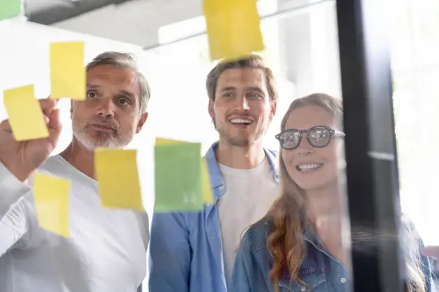 Three coworkers collaborating in an office using sticky notes on a glass wall.