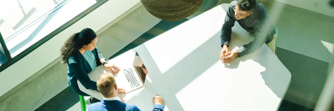 Three people are seated around a white table in a bright, modern office space, with one person using a laptop and sunlight streaming through large windows.