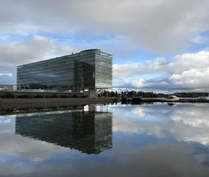 A modern glass office building stands by the waterfront, reflecting clearly on the calm water below. The structure has a sleek, rectangular design with multiple floors and large glass panels. The sky above is partly cloudy with patches of blue, and the surrounding area includes a few trees and distant buildings.
