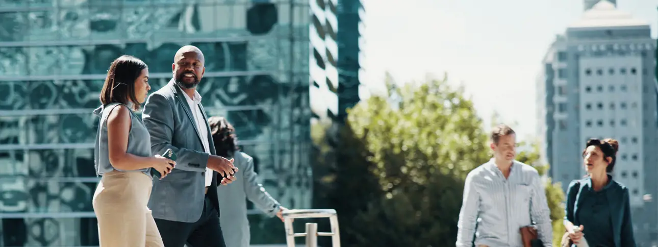 Alt text: Four people in business attire walking and talking outdoors in a city setting, with modern glass buildings and trees in the background.  