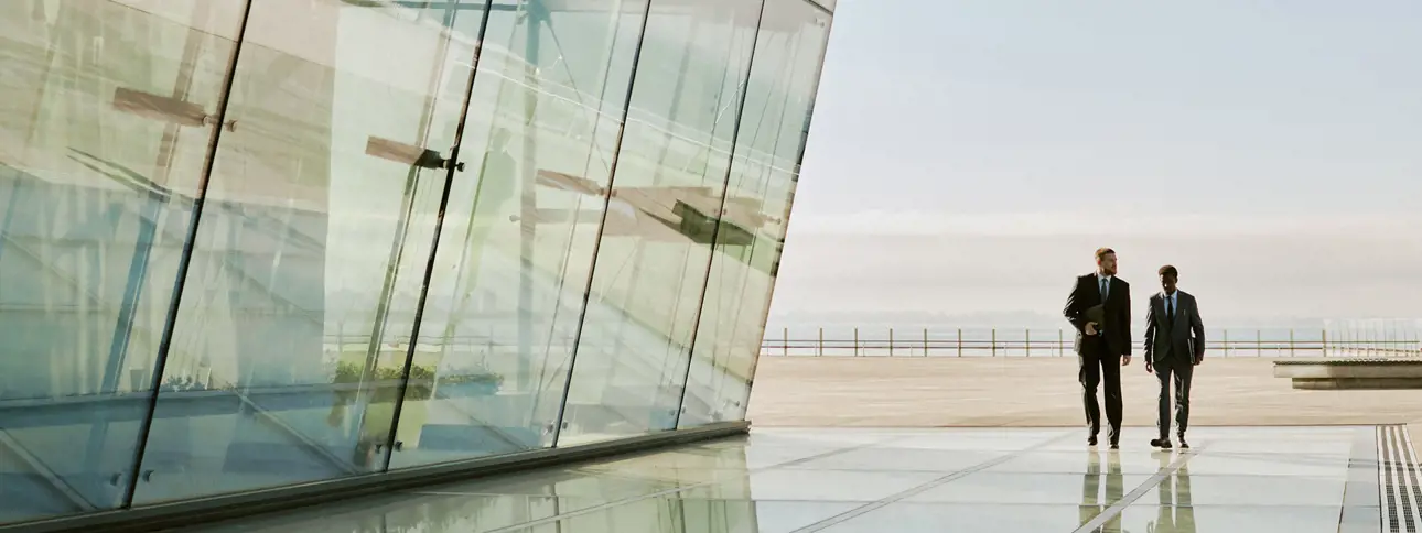Two people in business suits walking outside a modern glass building on a bright, clear day.