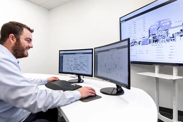 A man in a blue shirt smiling as he looks at a computer screen, seated at a white desk with white walls in the background.